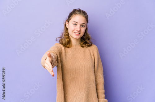 Young caucasian woman on purple background stretching hand at camera in greeting gesture.