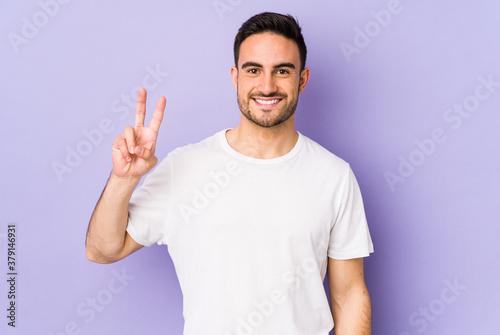 Young caucasian man isolated on purple background showing victory sign and smiling broadly.