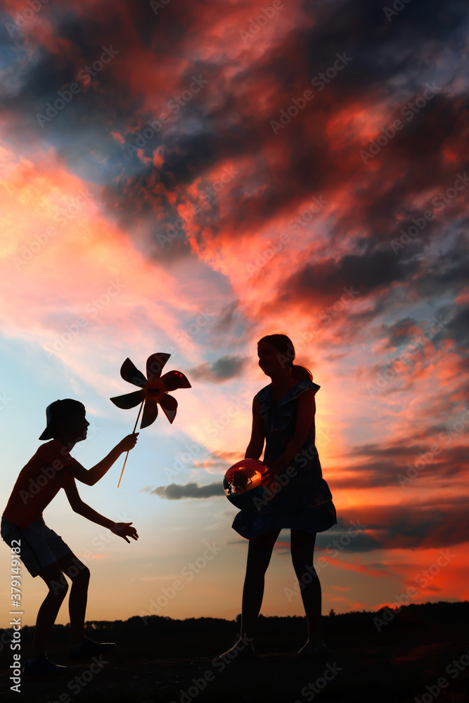 Silhouettes of playing boy with windmill and girl with ball in the nature in red blue dramatic cloudy sky background at sunset