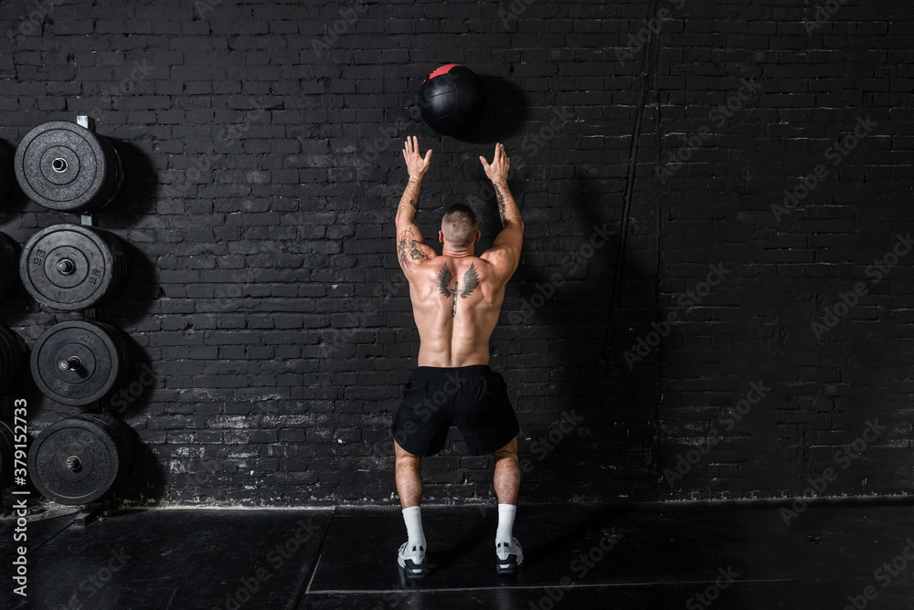 Young strong sweaty focused fit muscular man with big muscles doing throwing medicine ball up on the wall for cross training hard core workout in the gym real people