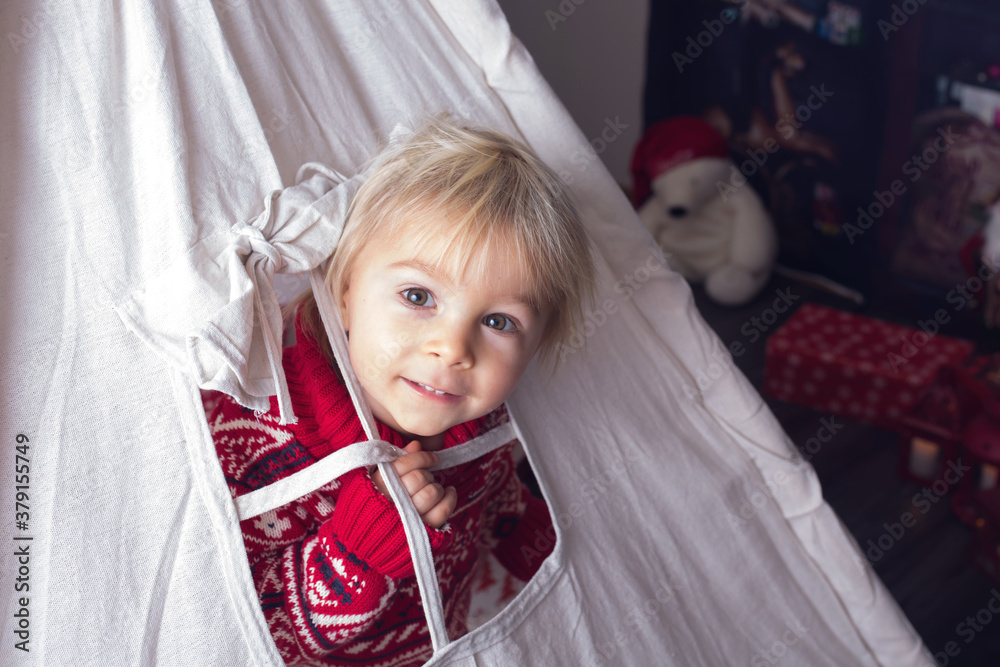 Cute little blonde boy with Christmas sweater, looking from a window of teepee at home Cute little blonde boy with Christmas sweater, looking from a window of teepee at home
