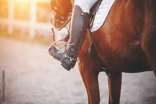 The foot of the rider sitting on the horse is dressed in a black boot with a spur and rests on a metal stirrup, illuminated by sunlight. Horseback riding. Equestrian sport.