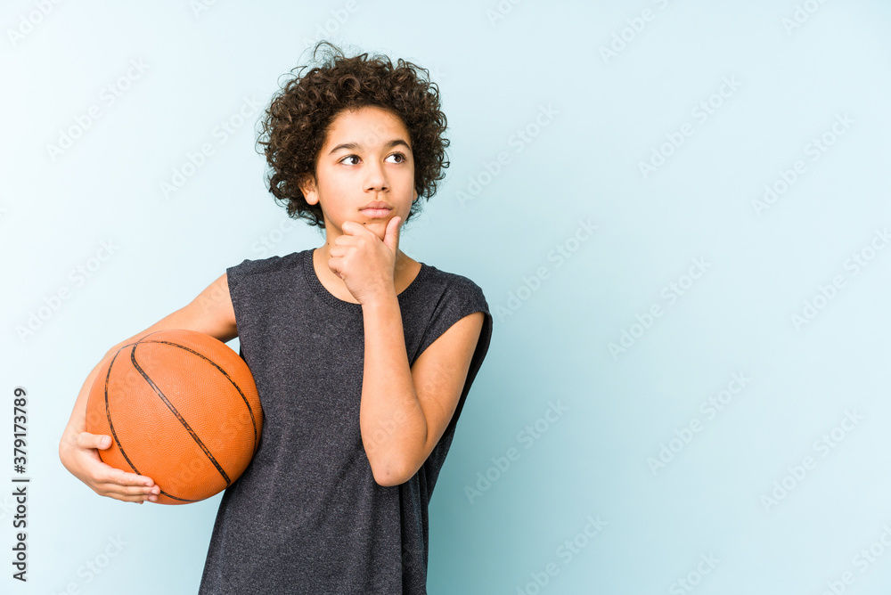 Kid boy playing basketball isolated on blue background looking sideways with doubtful and skeptical expression.