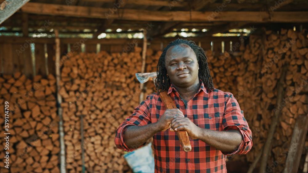 African man lumberjack with the axe standing in front of stack of cutted timber for winter. High quality photo