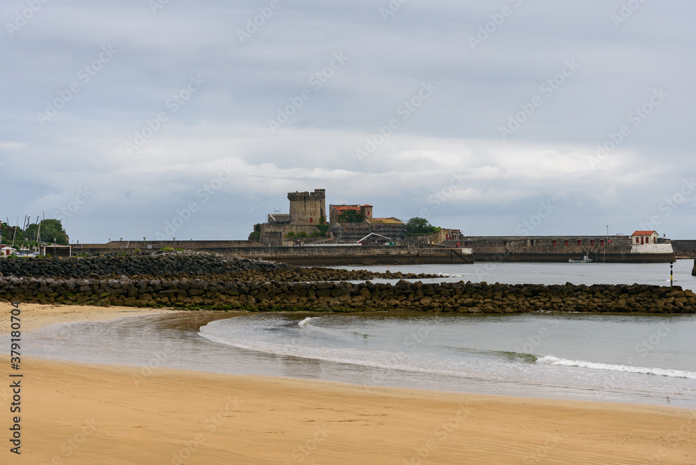 Close-up of socoa beach, at low tide, small waves in the middle of the rocks of the boardwalk at the mouth of the river. In the photo the castle of Sokoa above the gray clouds of the landscape