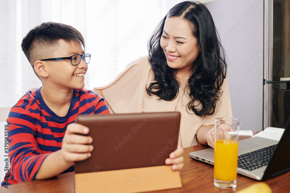 Proud smiling son showing tablet computer to motehr after finishing hard level in game Proud smiling son showing tablet computer to motehr after finishing hard level in game