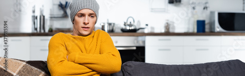 Fotografia horizontal crop of freezing man in warm sweater and hat sitting with crossed arm