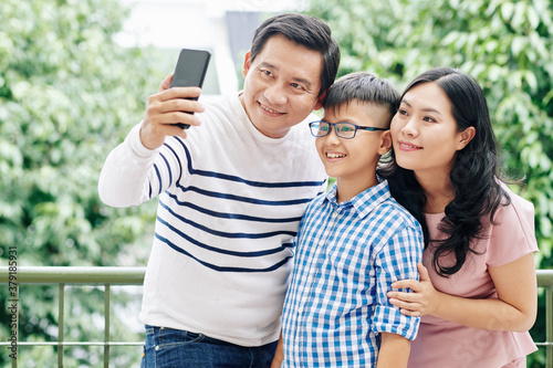 Mature Vietnamese man taking selfie with wife and preteen son on balcony