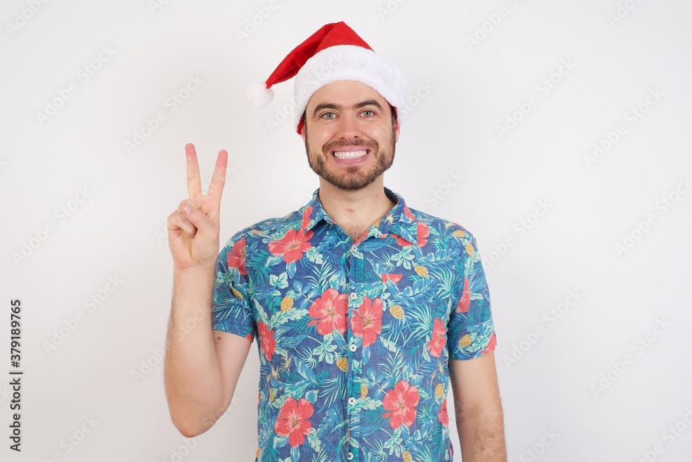 Young caucasian man wearing hawaiian shirt and Santa hat over isolated white background showing and pointing up with fingers number two while smiling confident and happy. Young caucasian man wearing hawaiian shirt and Santa hat over isolated white background showing and pointing up with fingers number two while smiling confident and happy.