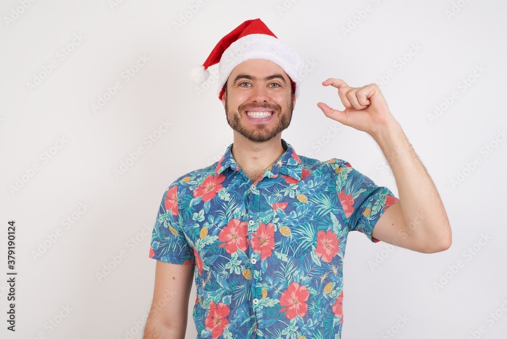 Young caucasian man wearing hawaiian shirt and Santa hat over isolated white backgr smiling and confident gesturing with hand doing small size sign with fingers looking and the camera. Measure concept