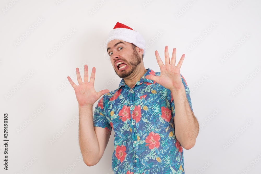 Young caucasian man wearing hawaiian shirt and Santa hat over isolated white background afraid and terrified with fear expression stop gesture with hands, shouting in shock. Panic concept.