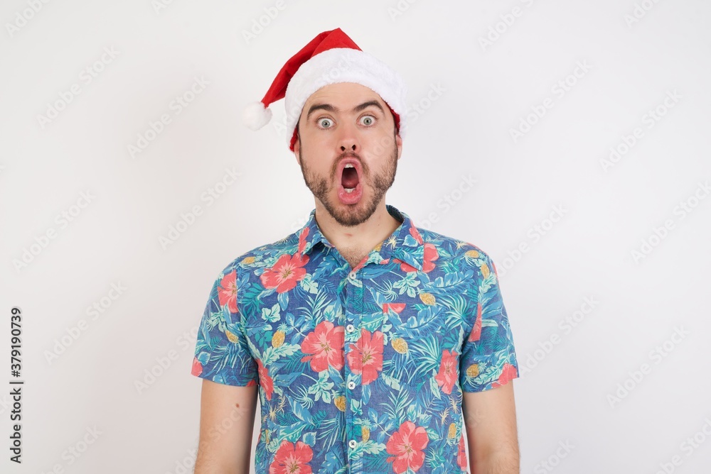 Oh my God. Surprised  Young caucasian man wearing hawaiian shirt and Santa hat over isolated white background stares at camera with shocked expression exclaims with unexpectedness,