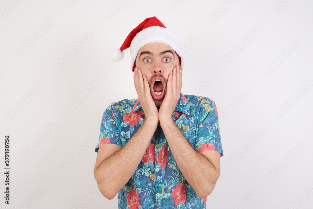 Scared terrified Young caucasian man wearing hawaiian shirt and Santa hat over isolated white wall shocked with prices at shop, being short of money to buy something, People and human emotions concept Scared terrified Young caucasian man wearing hawaiian shirt and Santa hat over isolated white wall shocked with prices at shop, being short of money to buy something, People and human emotions concept