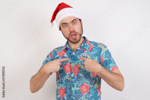 Photo of embarrassed Young caucasian man wearing hawaiian shirt and Santa hat over isolated white background indicates at hisself with puzzled expression, being shocked  photo