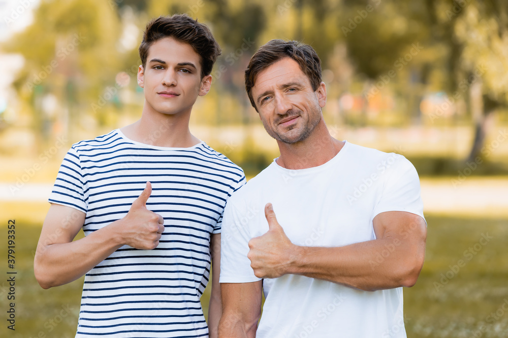 father and teenager son in t-shirts showing thumbs up in park father and teenager son in t-shirts showing thumbs up in park
