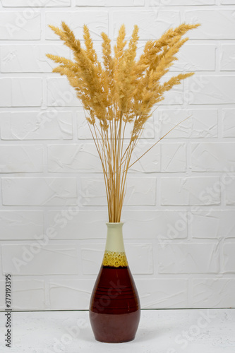 A bouquet of dry spikelets in a brown ceramic vase on a wooden table against a white brick background. Stylish home decor. Modern interior design photo