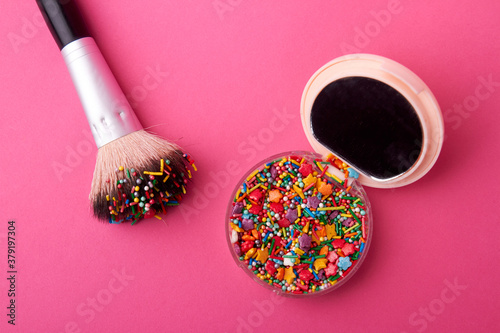 Cosmetic powder case with sweet sprinkles. Make-up brush with case with sweet candies isolated on pink background. photo