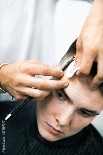 Selective focus of barber using sharp razor during haircut of young man in barbershop 