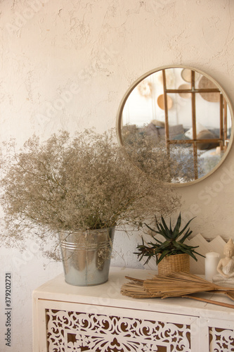 a bouquet of dried flowers in a bucket stands on a carved wooden white cabinet on the white wall background with mirror with boho style  interior in reflection