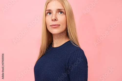 Portrait of gorgeous beautiful young blonde Caucasian female with freckles and blue eyes posing isolated in dark blue dress, looking up with thoughtful dreamy facial expression, making future plans