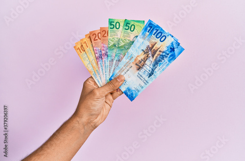 Hispanic hand holding swiss franc banknotes over isolated pink background.