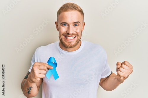 Young caucasian man holding blue ribbon screaming proud, celebrating victory and success very excited with raised arm