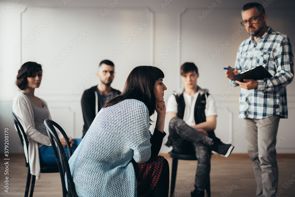 Handsome senior professional therapist stands in front of a group of people of different ages during meeting for people with social problems Handsome senior professional therapist stands in front of a group of people of different ages during meeting for people with social problems
