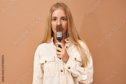 Horizontal shot of serious attractive young woman with long fair hair posing isolated with brush, going to paint walls in her new apartment. People, renovation, decoration and repair concept