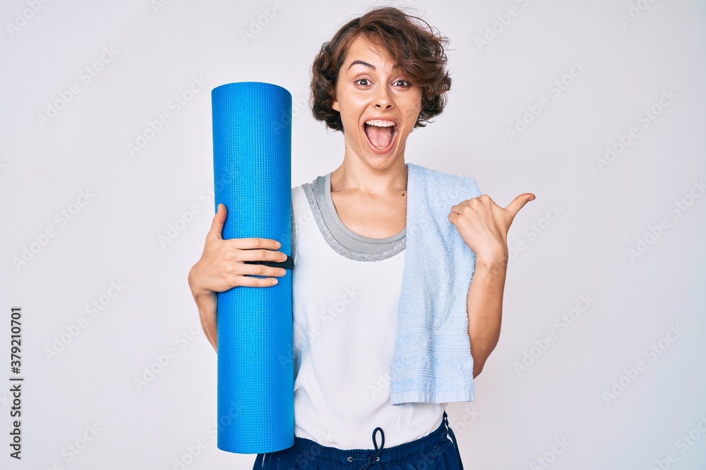 Young hispanic woman holding yoga mat pointing thumb up to the side smiling happy with open mouth Young hispanic woman holding yoga mat pointing thumb up to the side smiling happy with open mouth