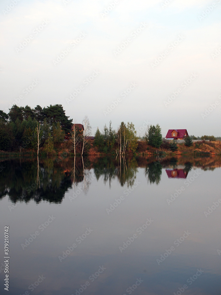 Autumn landscape with a lake surrounded by trees reflected in the water, against a blue sky. Rural landscape, a beautiful place to relax.