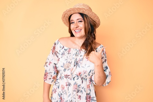 Young beautiful caucasian woman wearing summer dress and hat doing happy thumbs up gesture with hand. approving expression looking at the camera showing success.