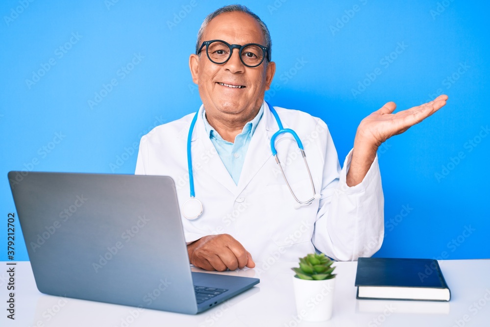 Senior handsome man with gray hair wearing doctor uniform working using computer laptop smiling cheerful presenting and pointing with palm of hand looking at the camera. Senior handsome man with gray hair wearing doctor uniform working using computer laptop smiling cheerful presenting and pointing with palm of hand looking at the camera.