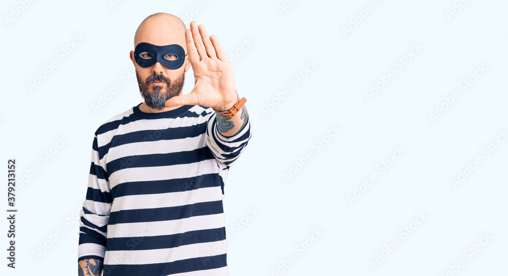 Young handsome man wearing burglar mask doing stop sing with palm of the hand. warning expression with negative and serious gesture on the face. Young handsome man wearing burglar mask doing stop sing with palm of the hand. warning expression with negative and serious gesture on the face.