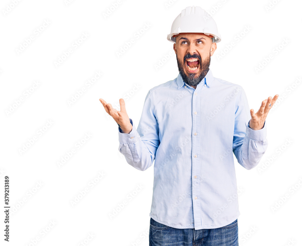 Young handsome man wearing architect hardhat crazy and mad shouting and yelling with aggressive expression and arms raised. frustration concept.