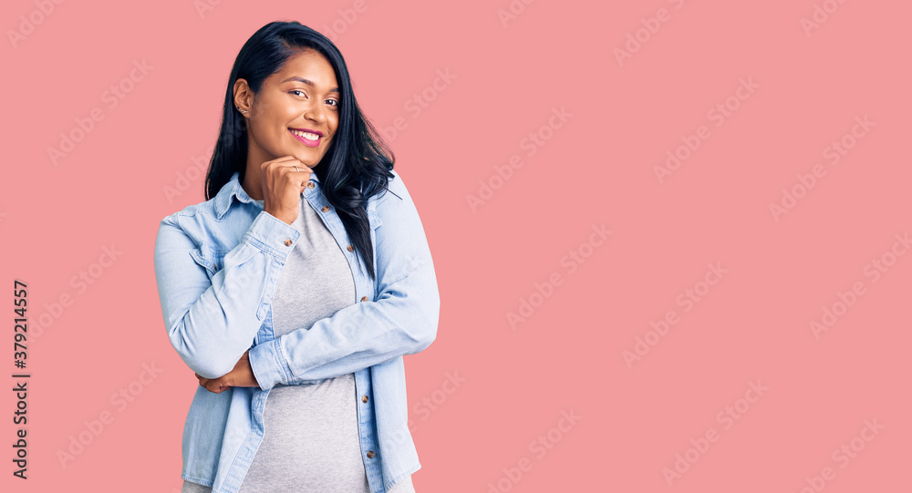 Hispanic woman with long hair wearing casual denim jacket looking confident at the camera with smile with crossed arms and hand raised on chin. thinking positive. Hispanic woman with long hair wearing casual denim jacket looking confident at the camera with smile with crossed arms and hand raised on chin. thinking positive.