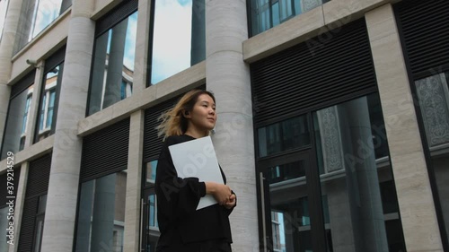 Attractive asian woman confidently walks through business district of metropolis, holding documents folder. Stylish female freelancer or businesswoman or office employee, concept, 4k slow motion