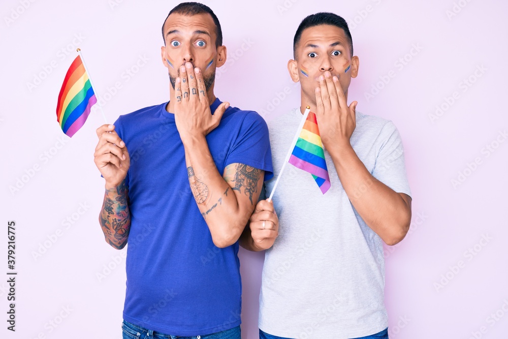 Young gay couple of two men holding rainbow lgbtq flags together covering mouth with hand, shocked and afraid for mistake. surprised expression