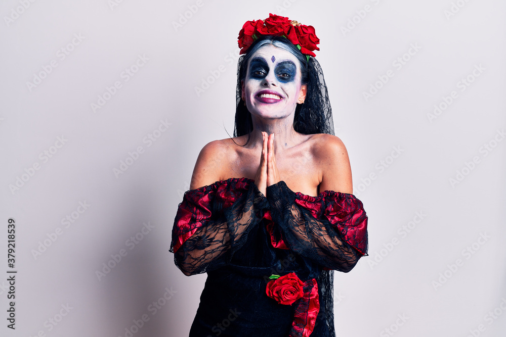 Young woman wearing mexican day of the dead makeup praying with hands together asking for forgiveness smiling confident.