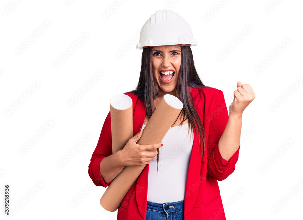 Young beautiful brunette woman wearing hardhat holding architect blueprint screaming proud, celebrating victory and success very excited with raised arms Young beautiful brunette woman wearing hardhat holding architect blueprint screaming proud, celebrating victory and success very excited with raised arms