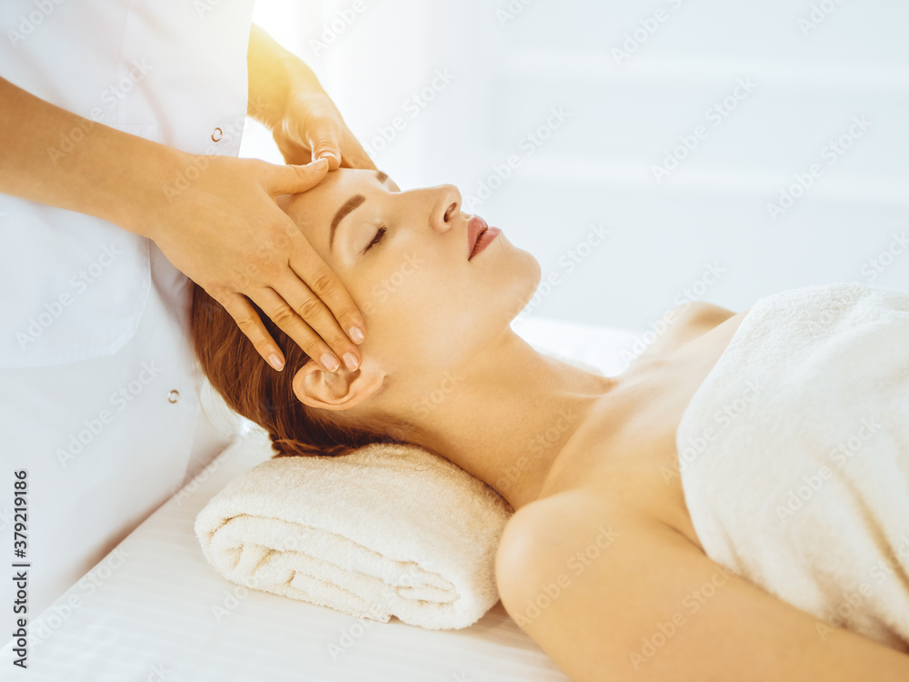Beautiful woman enjoying facial massage with closed eyes in sunny spa center. Relaxing treatment concept in medicine Beautiful woman enjoying facial massage with closed eyes in sunny spa center. Relaxing treatment concept in medicine