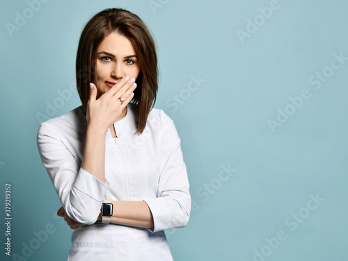 Shy positive woman dentist or doctor orthodontist in traditional white medical uniform standing and covering mouth with hand