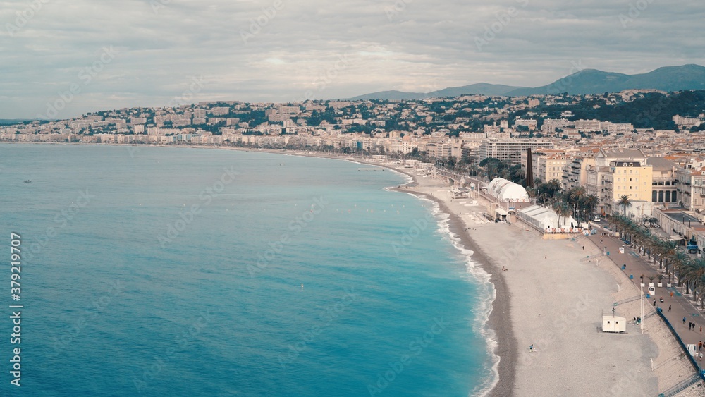 South city with a bright coastline in Nice, France, houses near a big ocean. Cloudy day, drone shot of long extended shoreline, cloudy windy day, sandy beach mountains on background