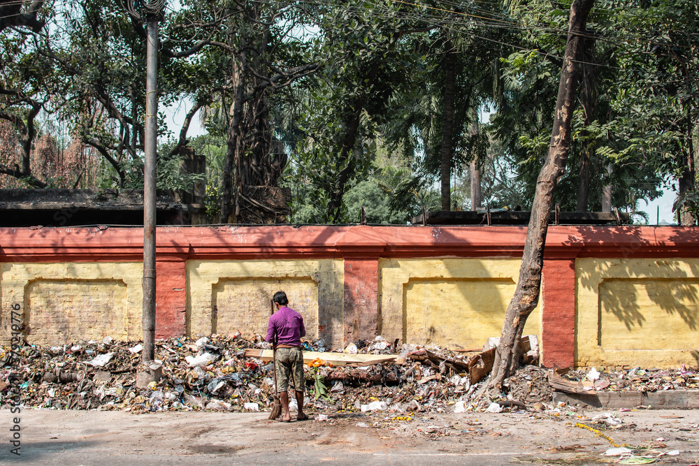 Fototapeta premium Kolkata, India - February 1, 2020: An unidentified man in purple shirt stands by himself with a broom infront of a big pile of plastic landfill waste on February 1, 2020 in Kolkata, India