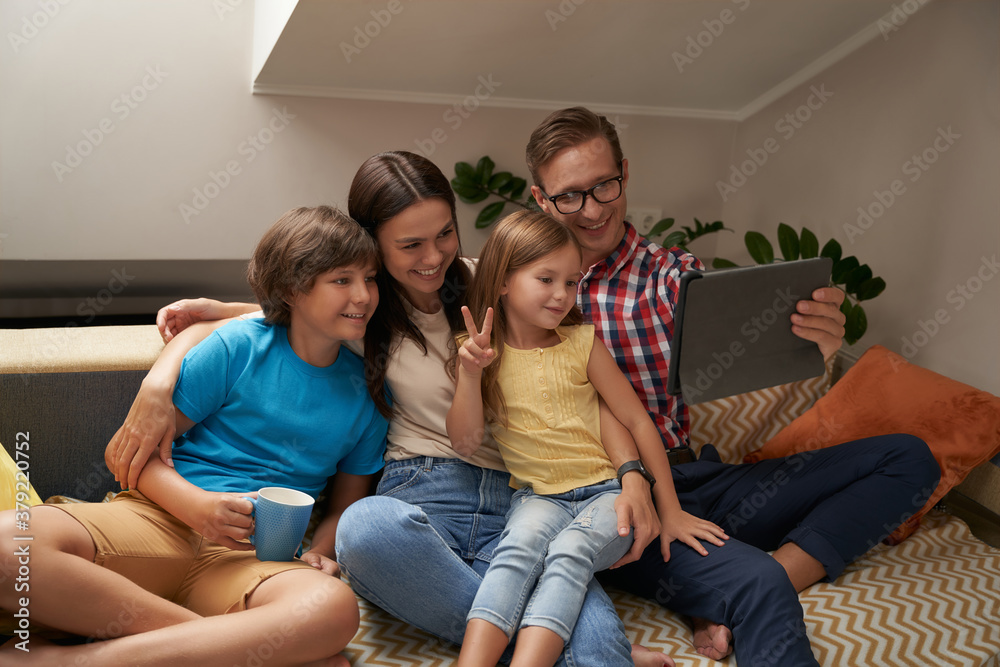 Parents and their two little kids sitting on sofa in the living room and using digital tablet, they looking at web camera and smiling while having video call