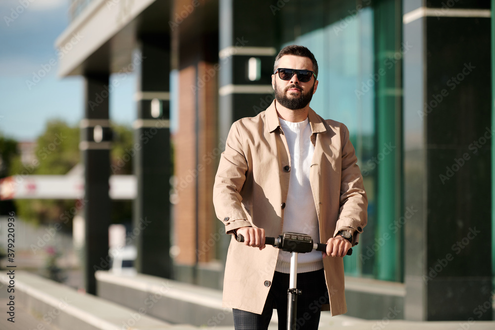 Serious bearded businessman in beige trenchcoat and sunglasses riding scooter Serious bearded businessman in beige trenchcoat and sunglasses riding scooter