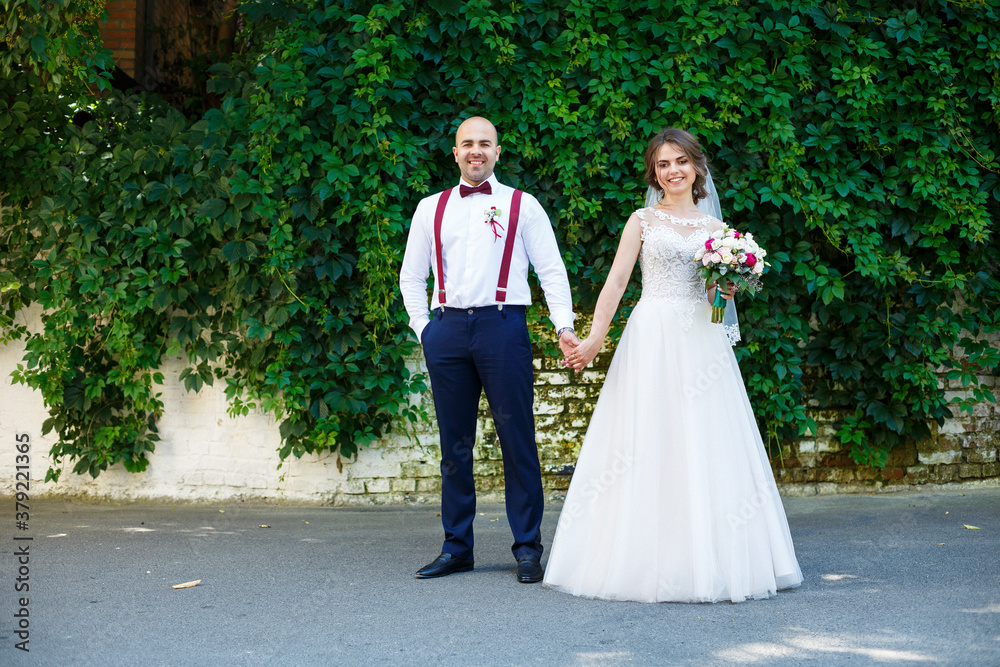 Beautiful couple bride in a white dress with a bouquet while the groom with suspenders and bow tie holding hands. Against the background of a wall with green leaves. Happy couple. Marriage concept. Beautiful couple bride in a white dress with a bouquet while the groom with suspenders and bow tie holding hands. Against the background of a wall with green leaves. Happy couple. Marriage concept.