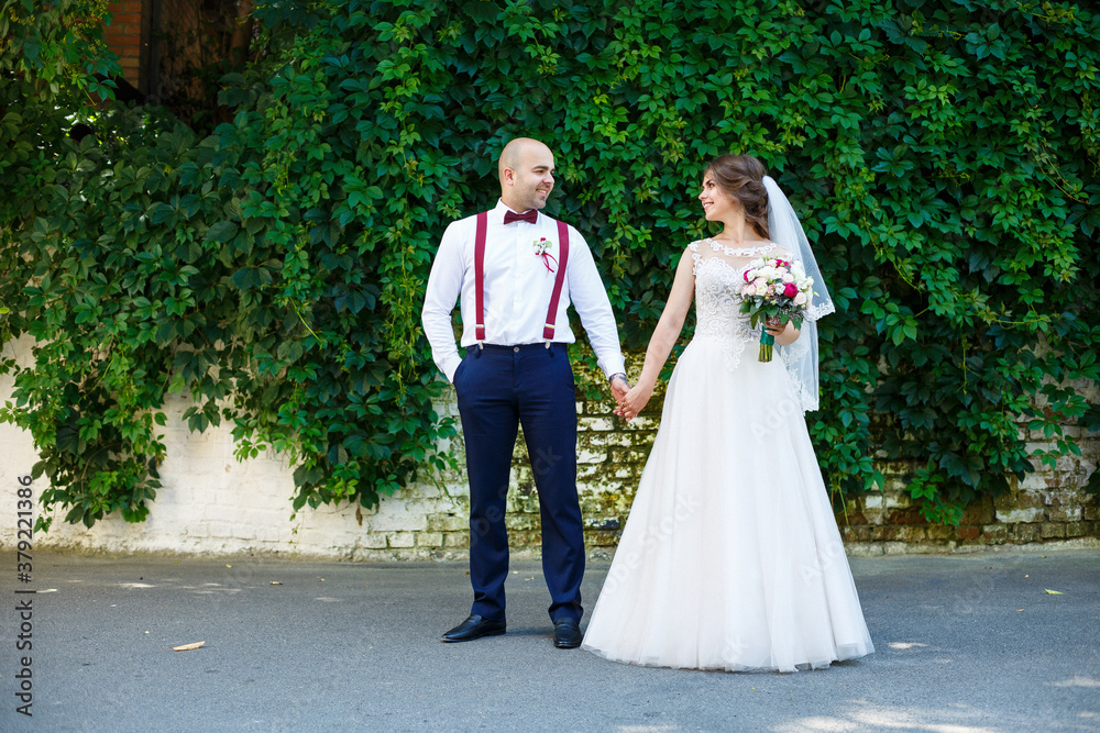 Beautiful couple bride in a white dress with a bouquet while the groom with suspenders and bow tie holding hands. Against the background of a wall with green leaves. Happy couple. Marriage concept. Beautiful couple bride in a white dress with a bouquet while the groom with suspenders and bow tie holding hands. Against the background of a wall with green leaves. Happy couple. Marriage concept.