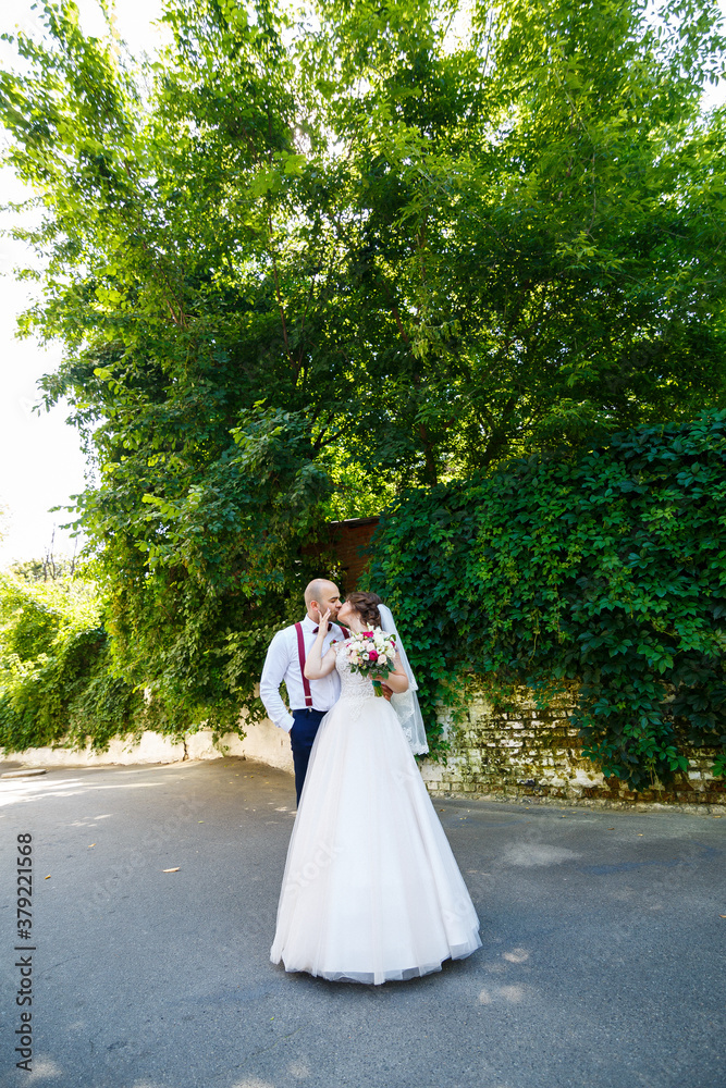 Beautiful couple bride in a white dress with a bouquet while the groom with suspenders and bow tie holding hands. Against the background of a wall with green leaves. Happy couple. Marriage concept. Beautiful couple bride in a white dress with a bouquet while the groom with suspenders and bow tie holding hands. Against the background of a wall with green leaves. Happy couple. Marriage concept.