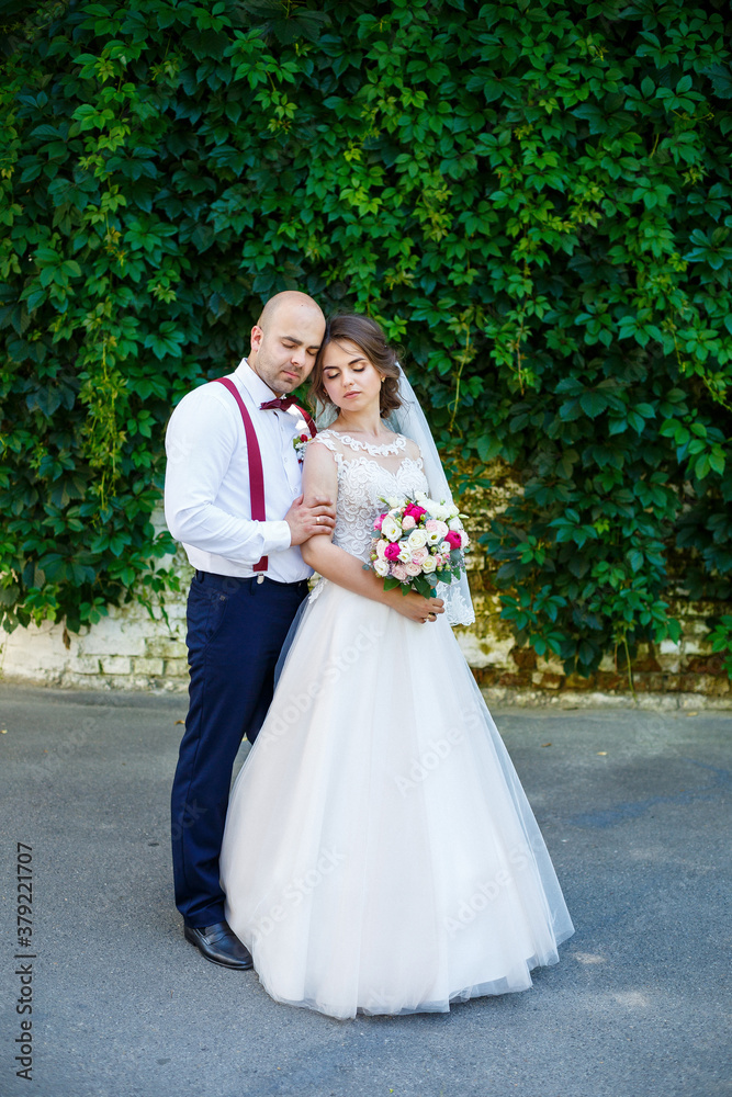Cheerful couple bride in a white dress with a bouquet while the groom with suspenders and bow tie. Against the background of a wall with green leaves. Happy couple. The concept of marriage.