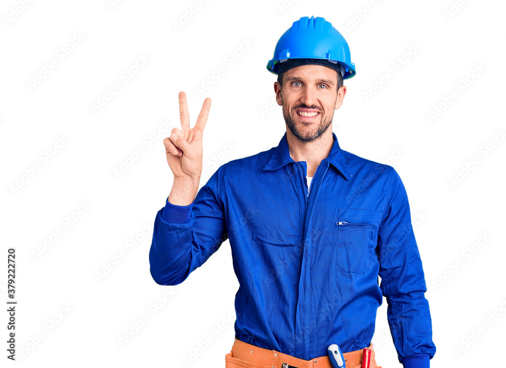 Young handsome man wearing worker uniform and hardhat smiling looking to the camera showing fingers doing victory sign. number two.